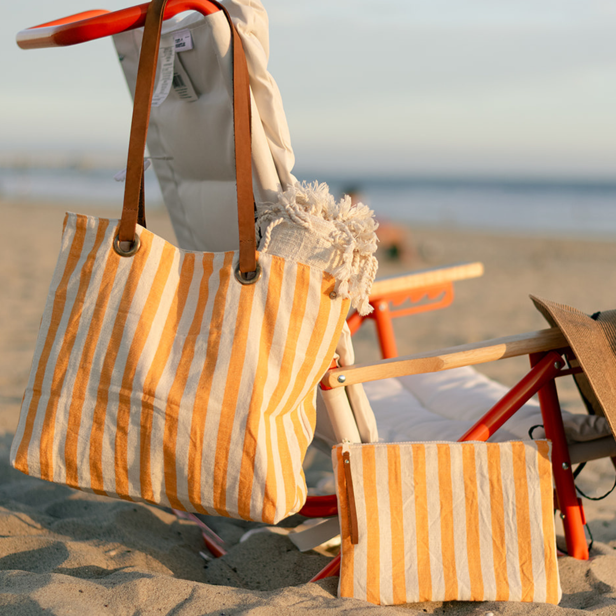 A striped beach tote bag and matching pouch with yellow vertical stripes, hanging on a beach chair at the beach.