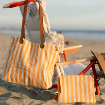 A striped beach tote bag and matching pouch with yellow vertical stripes, hanging on a beach chair at the beach.