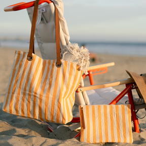 A striped beach tote bag and matching pouch with yellow vertical stripes, hanging on a beach chair at the beach.